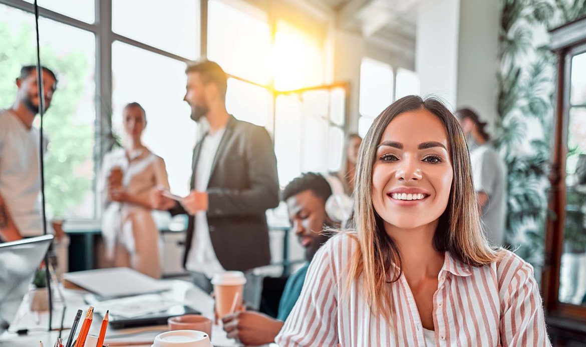 Smiling woman in a bustling office environment, embodying resilience and the ability to reframe thinking by adapting in the face of challenges.