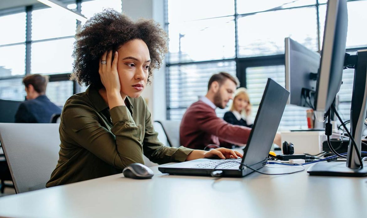 Young woman appearing distracted and unfocused at her desk in a busy office, illustrating common signs of ADHD in a work environment.