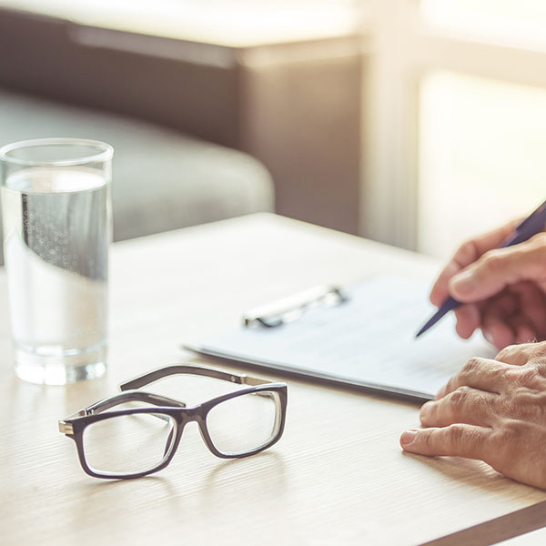 A close-up of a hand writing on a clipboard with glasses and a glass of water on a table, representing Schizoaffective Disorder symptoms.