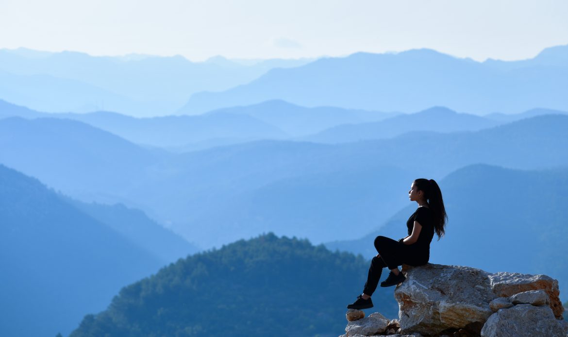 Woman sitting alone on a mountain cliff, gazing thoughtfully into the distance, reflecting the isolating and contemplative effects of negative thinking.