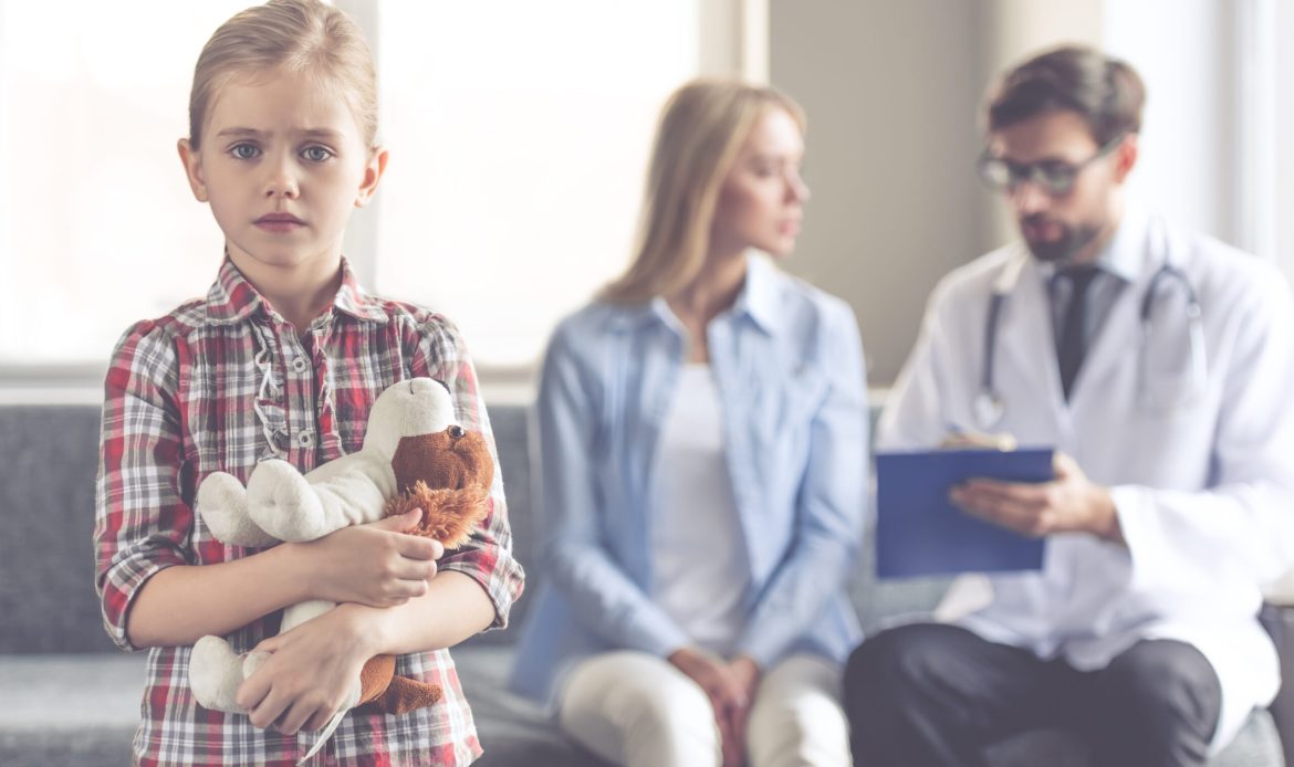 Beautiful young mother and her little daughter at the pediatrician. Girl is holding a toy and looking sadly at camera