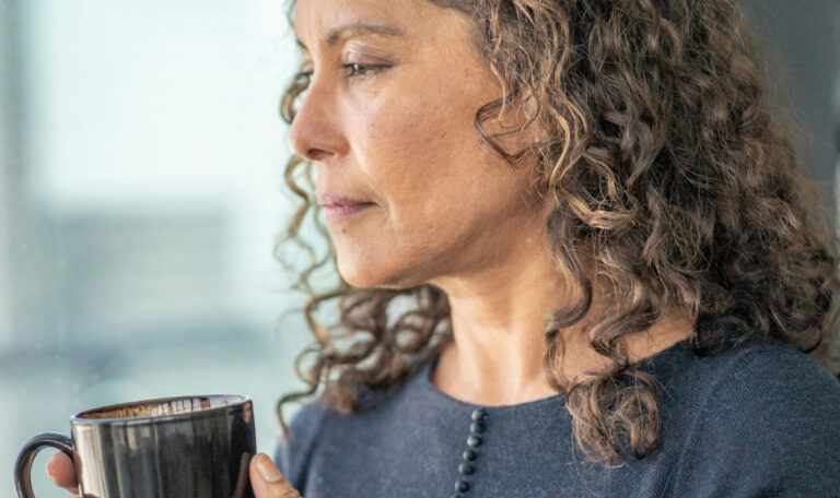 Woman gazing thoughtfully out a window, holding a coffee cup, capturing a quiet moment filled with both joy and pain.