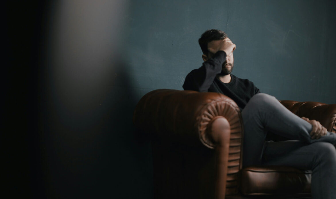 A person sits on a couch with their hand on their forehead, suggesting psychological symptoms related to situational anxiety.