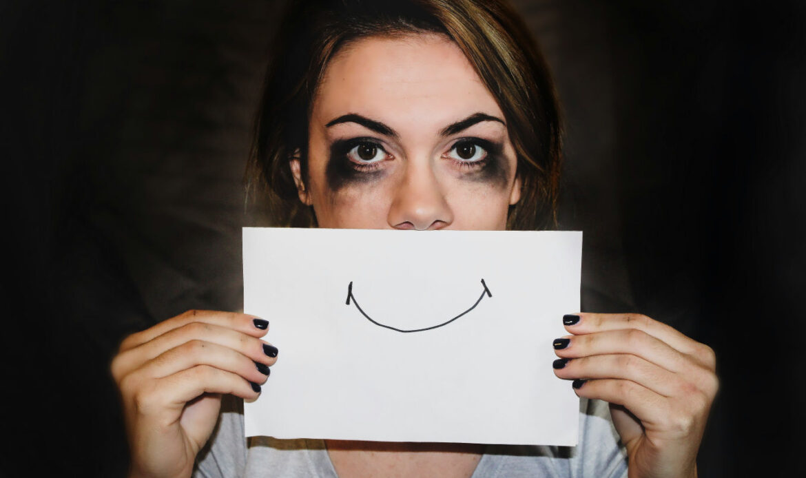 A woman holds a paper with a drawn smile in front of their mouth, with smudged makeup around their eyes, symbolizing situational anxiety.