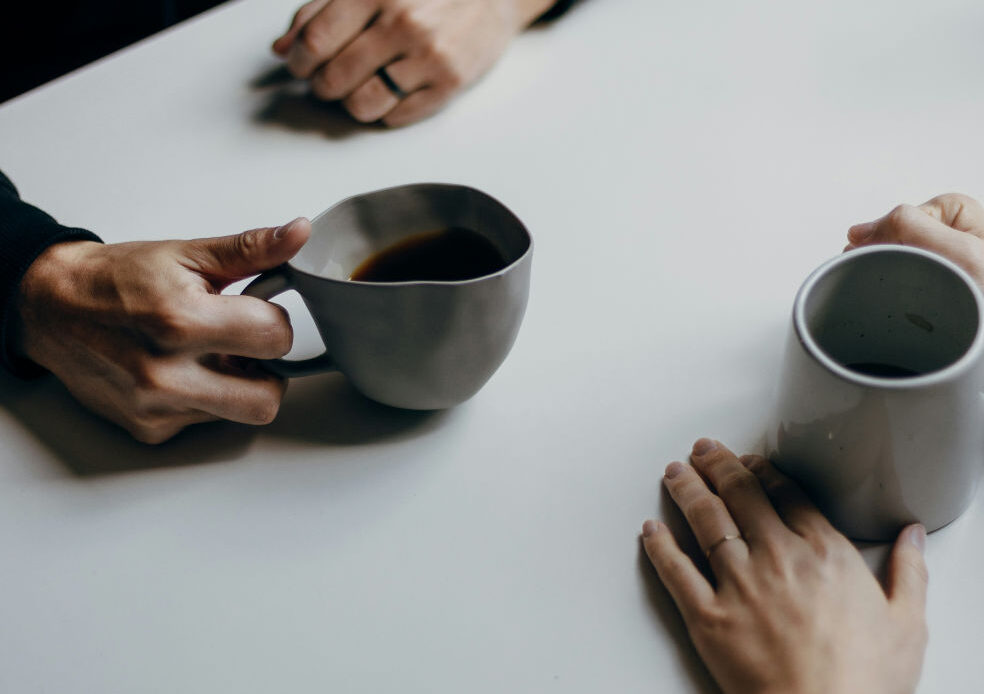 Two people sit across from each other at a table with coffee mugs, suggesting a conversation about seeking professional help for situational anxiety.