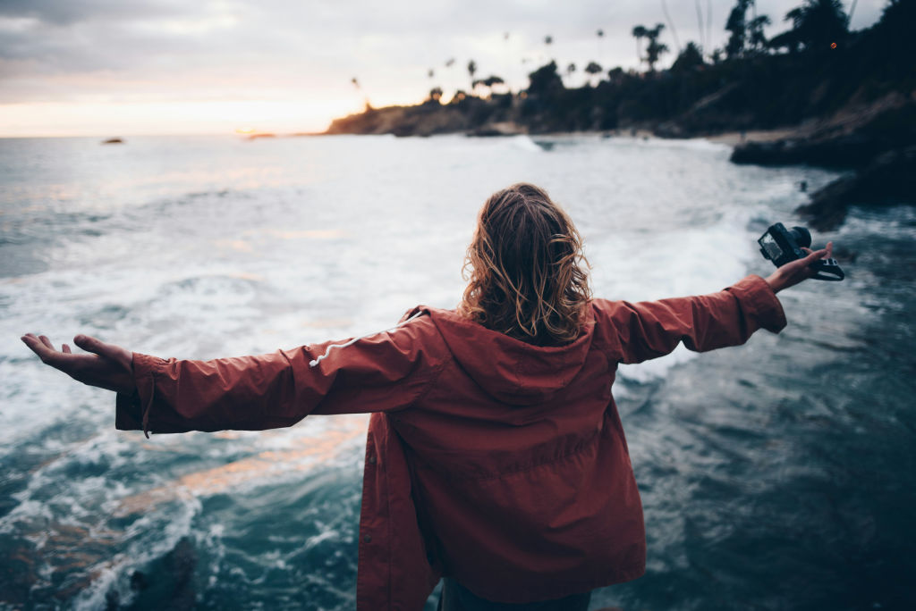 A person stands on a rocky shoreline with arms outstretched, facing the ocean waves, symbolizing awareness and acceptance of the present moment.