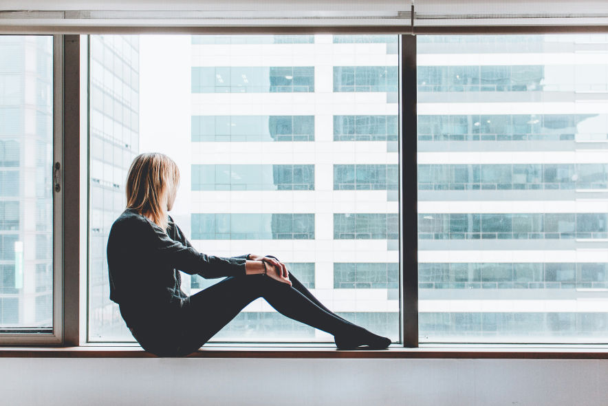 A woman sitting on a window ledge, gazing outside with a contemplative expression, symbolizing reflection and allowing oneself to grieve on the one-year death anniversary.