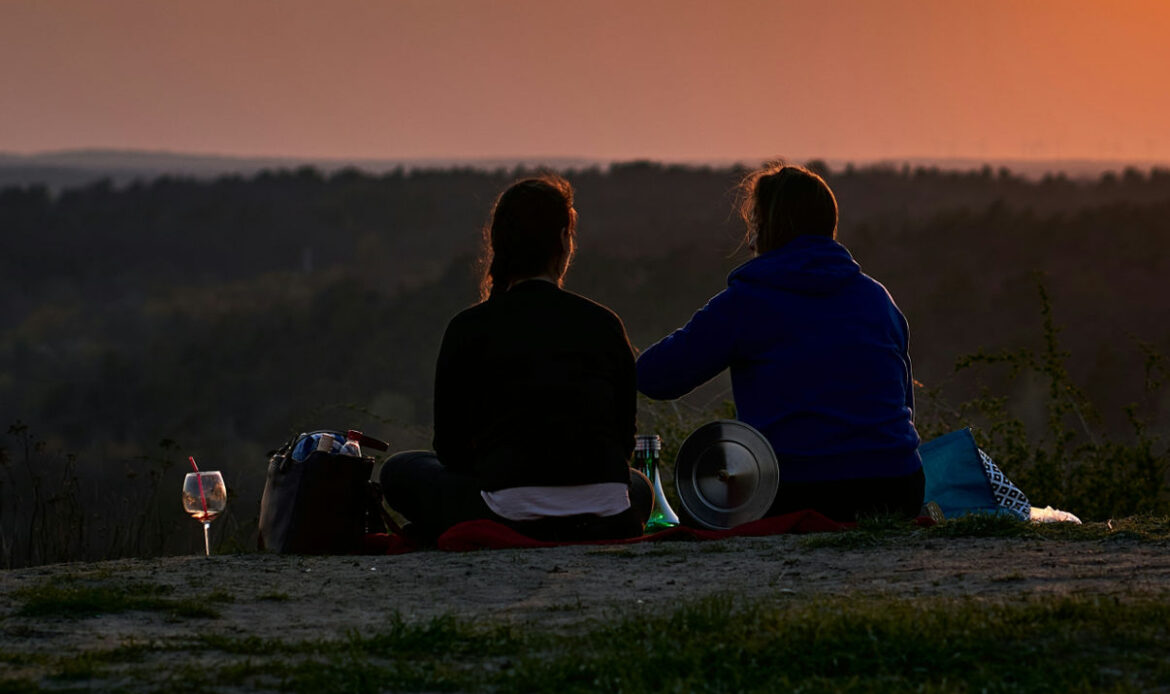 Two women sit together on a blanket overlooking a sunset, symbolizing being present, offering silent support, and providing companionship to a woman grieving the loss of her husband.