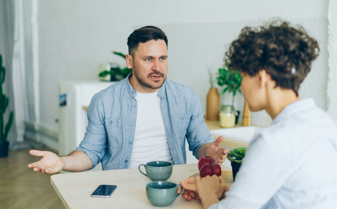  A man and woman sit at a table in a tense conversation, with the man appearing frustrated, symbolizing the emotional weight of feeling like a burden in a toxic relationship.