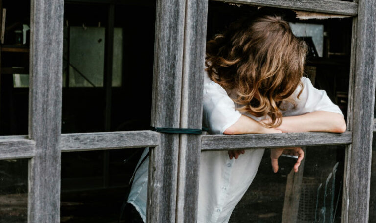 A woman leans out of a broken window with her head down, symbolizing isolation, overwhelm, and the weight of feeling like a burden.