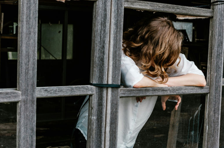 A woman leans out of a broken window with her head down, symbolizing isolation, overwhelm, and the weight of feeling like a burden.