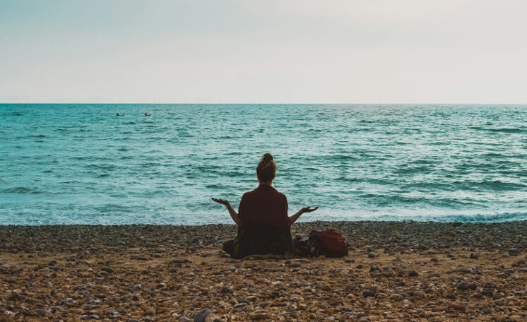 A person sits cross-legged on a rocky beach facing the ocean, arms open in a meditative pose, symbolizing peace and reflection during a healing journey.