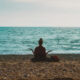 A person sits cross-legged on a rocky beach facing the ocean, arms open in a meditative pose, symbolizing peace and reflection during a healing journey.