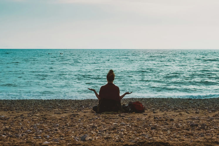 A person sits cross-legged on a rocky beach facing the ocean, arms open in a meditative pose, symbolizing peace and reflection during a healing journey.