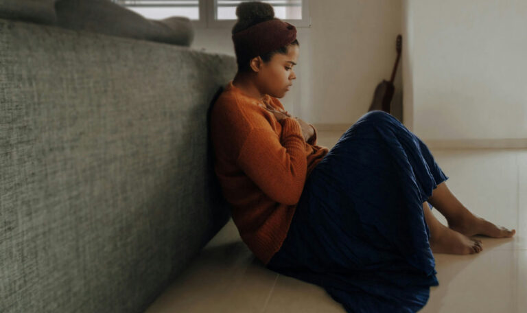 A woman sits on the floor with her back against a couch, holding her chest and looking inward, representing the process of self-reflection and learning how to heal anxious attachment.