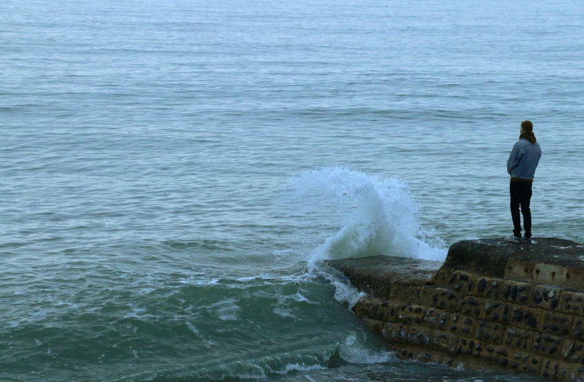 A person standing on a rocky pier, gazing at the ocean as waves crash against the shore, symbolizing the process of building a life around grief.