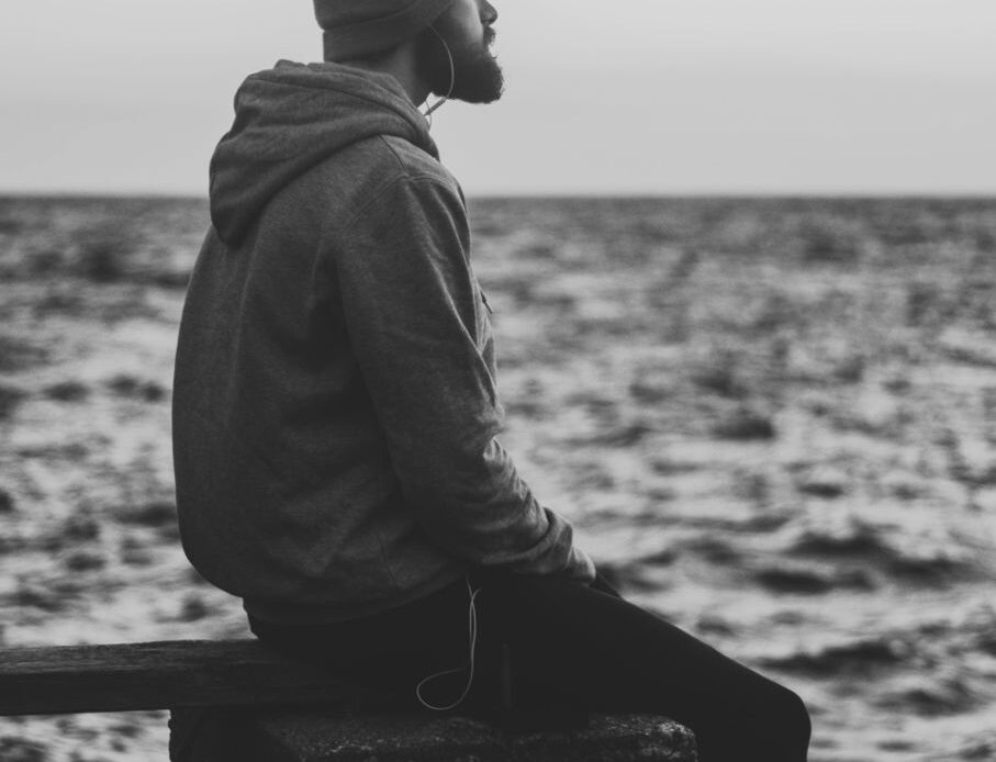 A person sitting alone by the water, wearing a hoodie and beanie, appearing deep in thought, symbolizing withdrawal from relationships or activities due to unresolved grief."