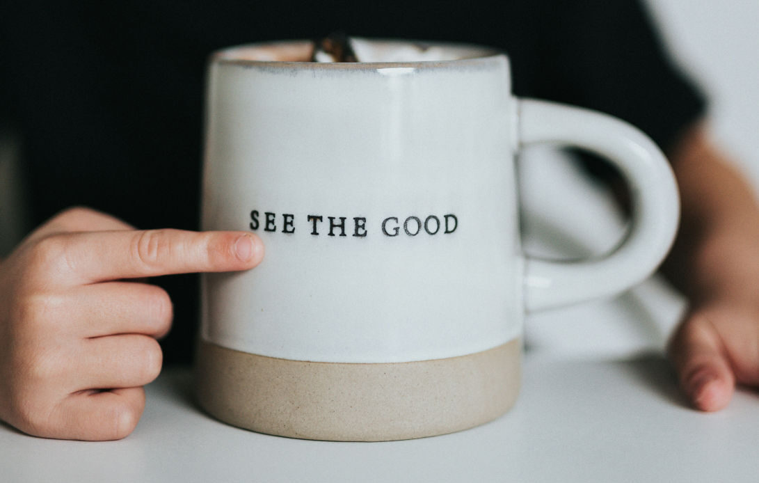 A person pointing to a white coffee mug with the phrase 'See The Good' printed on it, symbolizing a reminder for practicing positive affirmations.