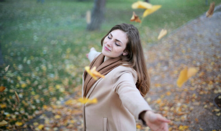 A woman standing in a park with her arms outstretched, eyes closed, and surrounded by falling autumn leaves, symbolizing peace and healing affirmations.