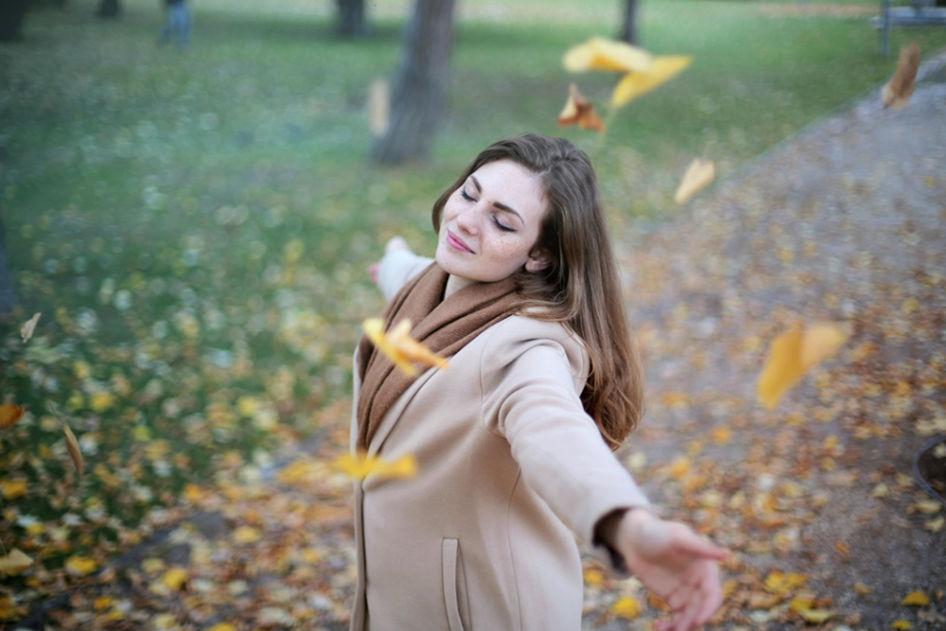 A woman standing in a park with her arms outstretched, eyes closed, and surrounded by falling autumn leaves, symbolizing peace and healing affirmations.