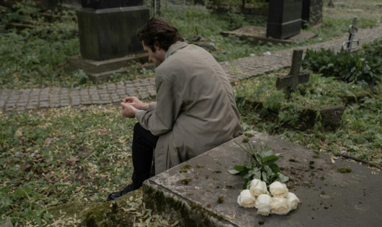 A person sitting by a grave in a cemetery, deep in thought, with white roses placed on a tombstone, symbolizing unresolved grief.