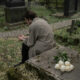 A person sitting by a grave in a cemetery, deep in thought, with white roses placed on a tombstone, symbolizing unresolved grief.