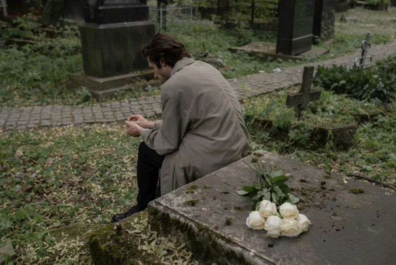 A person sitting by a grave in a cemetery, deep in thought, with white roses placed on a tombstone, symbolizing unresolved grief.