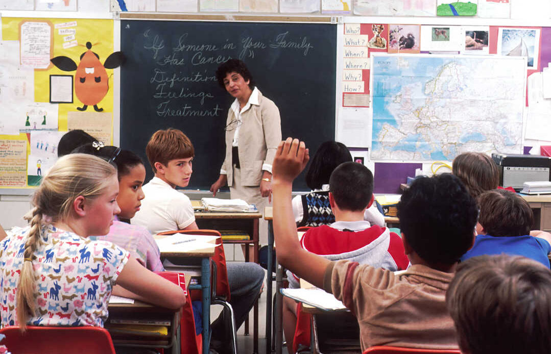 A classroom scene where a student raises their hand while others listen to a teacher, symbolizing active participation and the spirit of volunteering or giving back.